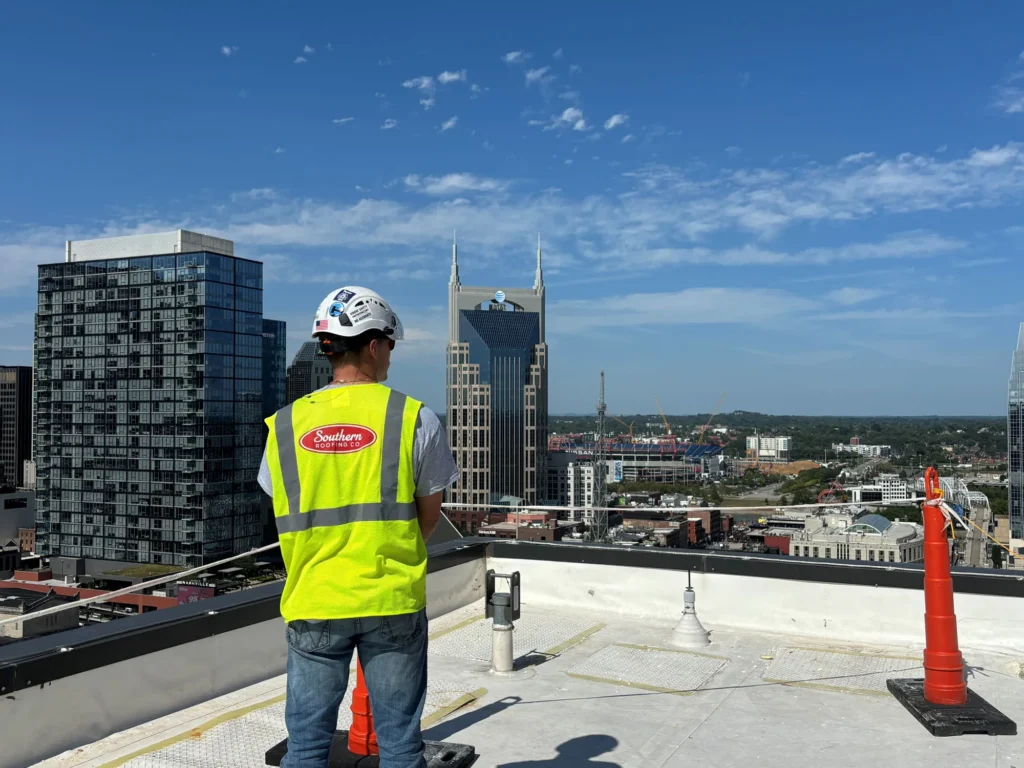 Southern Roofing Co worker in a safety vest standing on a downtown Nashville commercial rooftop facing the AT&T Building and skyline